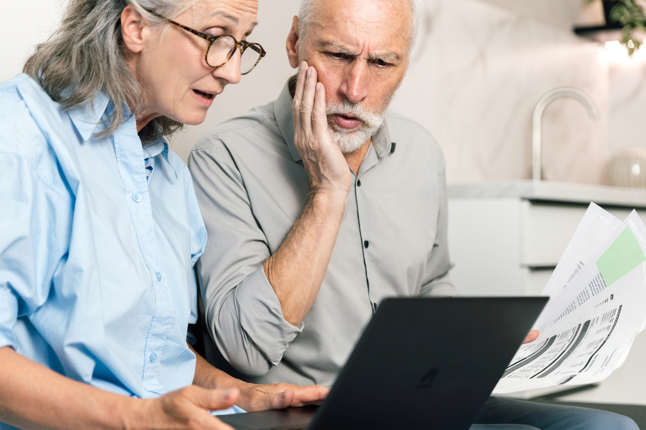 A concerned senior couple sitting at a desk looking at a laptop and financial bills.