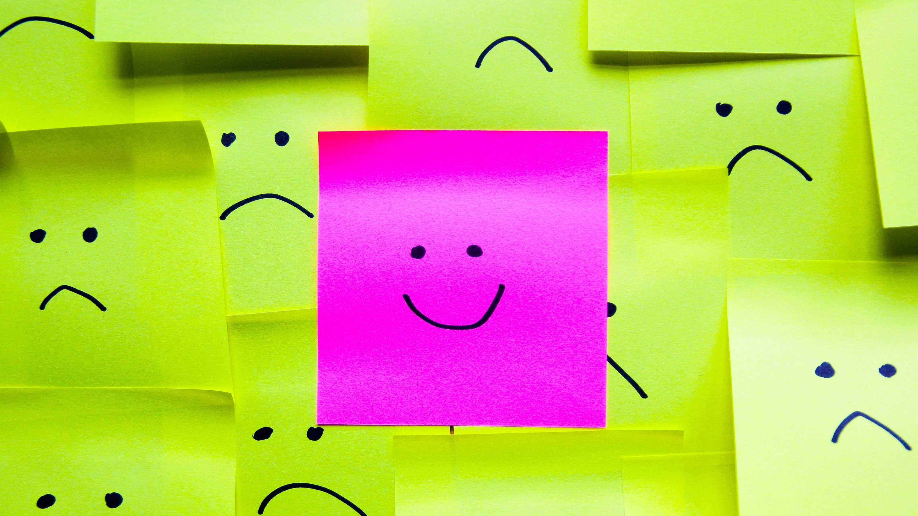 A pink post-it note with a smiley face standing out among several yellow post-it notes with sad faces.