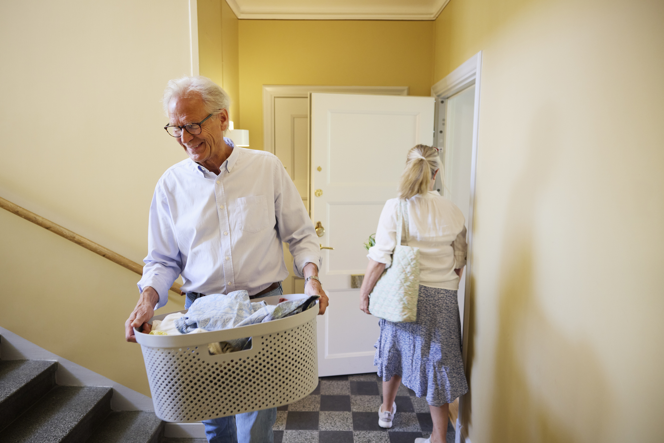 A mature man carrying a full laundry basket while passing his wife in a domestic setting.