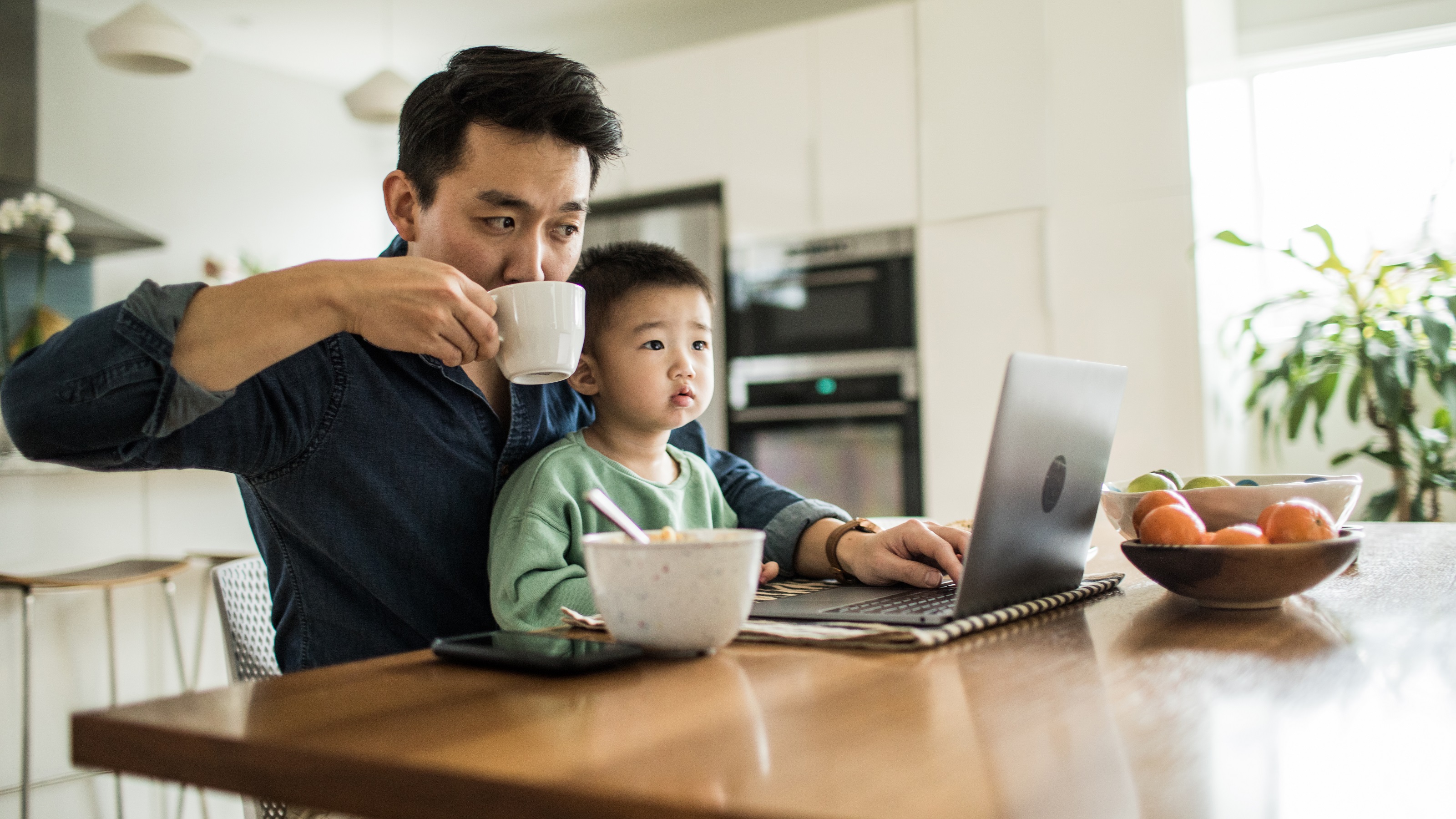 A father managing household finances on a laptop with a young child on his lap.