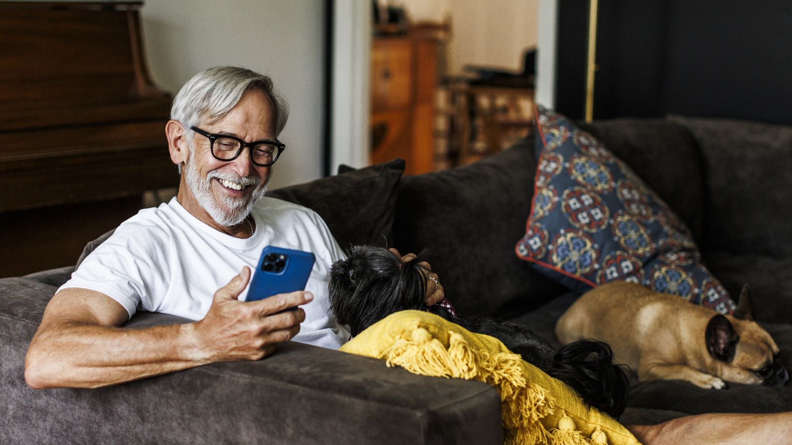 A senior man relaxing on a sofa at home with his two dogs, reading something on his smartphone.