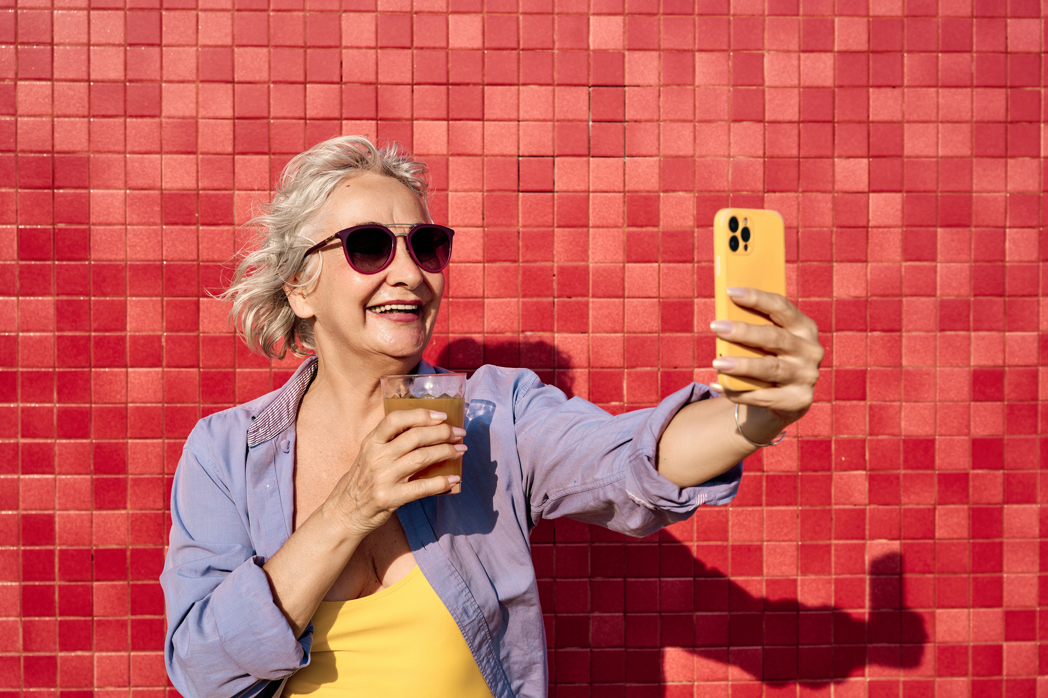 An energetic older woman taking a selfie with a drink in front of a vibrant red wall.